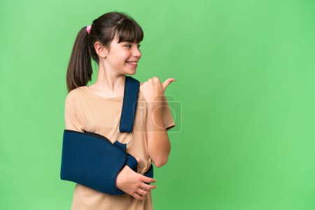 Photo for Little caucasian girl with broken arm and wearing a sling over isolated background pointing to the side to present a product - Royalty Free Image