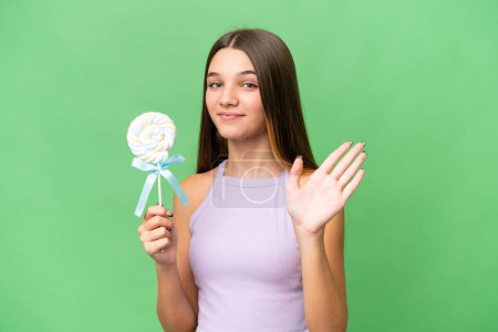 Foto de Adolescente chica caucásica sosteniendo una piruleta sobre un fondo aislado saludando con la mano con expresión feliz - Imagen libre de derechos