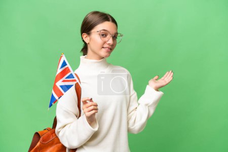 Téléchargez les photos : Adolescente étudiante caucasienne tenant un drapeau du Royaume-Uni sur fond isolé étendant les mains sur le côté pour inviter à venir - en image libre de droit