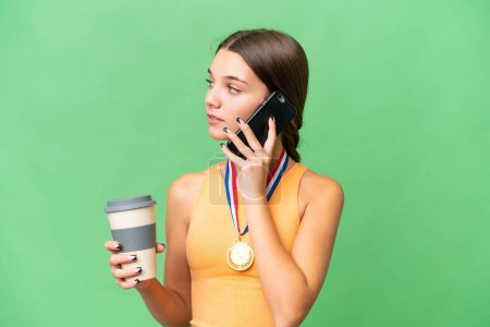 Photo for Teenager caucasian girl with medals over isolated background holding coffee to take away and a mobile - Royalty Free Image
