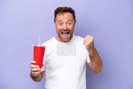 Foto de Hombre caucásico de mediana edad sosteniendo soda aislada sobre fondo púrpura celebrando una victoria en posición ganadora - Imagen libre de derechos