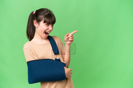 Photo for Little caucasian girl with broken arm and wearing a sling over isolated background pointing finger to the side and presenting a product - Royalty Free Image