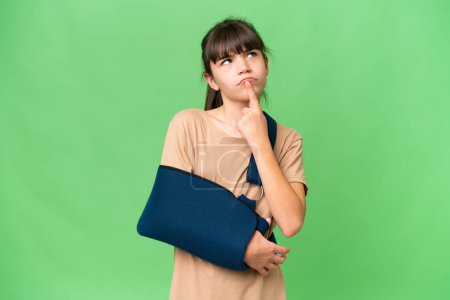 Photo for Little caucasian girl with broken arm and wearing a sling over isolated background having doubts while looking up - Royalty Free Image