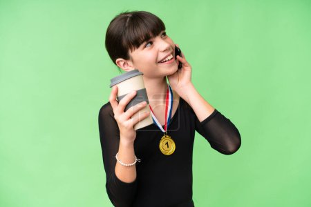 Photo for Little caucasian girl with medals over isolated background holding coffee to take away and a mobile - Royalty Free Image