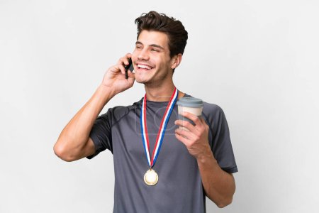 Photo for Young caucasian man with medals over isolated white background holding coffee to take away and a mobile - Royalty Free Image