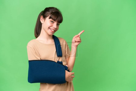 Photo for Little caucasian girl with broken arm and wearing a sling over isolated background pointing finger to the side - Royalty Free Image