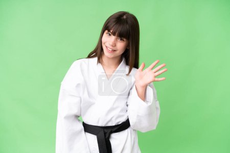 Kleines kaukasisches Mädchen beim Karate vor isoliertem Hintergrund salutiert mit glücklicher Miene mit der Hand