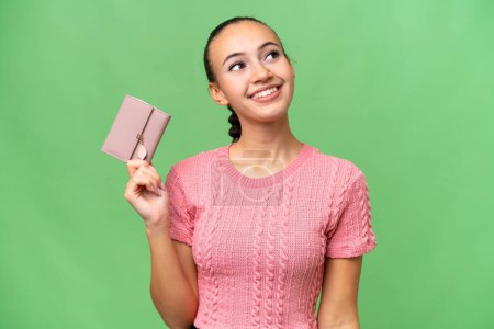 Photo for Young Arab woman holding a wallet over isolated background looking up while smiling - Royalty Free Image