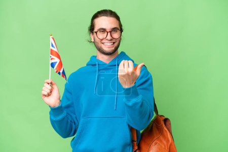 Téléchargez les photos : Jeune homme beau tenant un drapeau du Royaume-Uni sur fond isolé invitant à venir avec la main. Heureux que tu sois venu - en image libre de droit