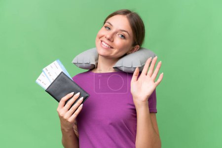 Young beautiful blonde woman with Inflatable travel Pillow over isolated background saluting with hand with happy expression