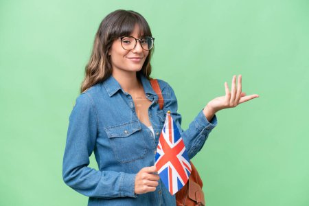 Jeune femme caucasienne tenant un drapeau du Royaume-Uni sur fond isolé étendant les mains sur le côté pour inviter à venir