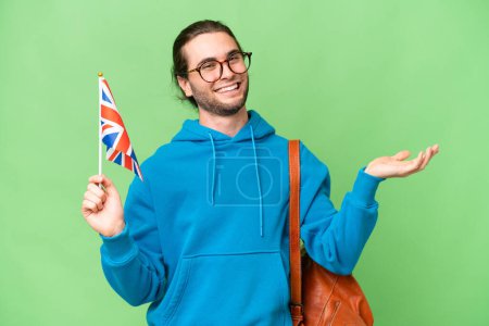 Téléchargez les photos : Jeune homme beau tenant un drapeau du Royaume-Uni sur fond isolé étendant les mains sur le côté pour inviter à venir - en image libre de droit
