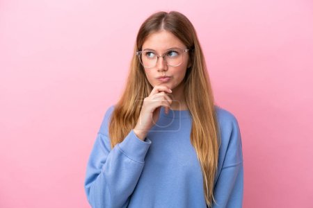 Young blonde woman isolated on pink background With glasses and having doubts