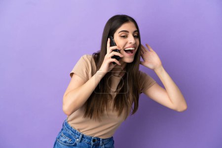 Photo for Young caucasian woman using mobile phone isolated on purple background listening to something by putting hand on the ear - Royalty Free Image
