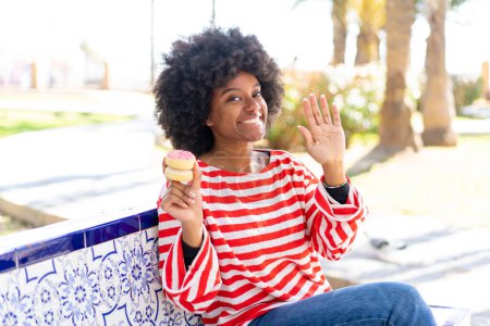 Foto de Chica afroamericana sosteniendo una dona al aire libre saludando con la mano con expresión feliz - Imagen libre de derechos