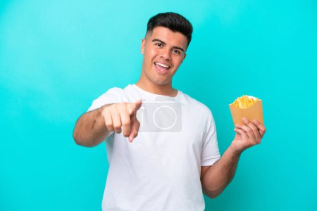 Foto de Joven caucásico hombre la captura de papas fritas aisladas sobre fondo azul sorprendido y apuntando frente - Imagen libre de derechos