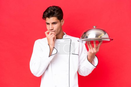 Photo for Young caucasian chef man holding tray isolated on red background having doubts - Royalty Free Image