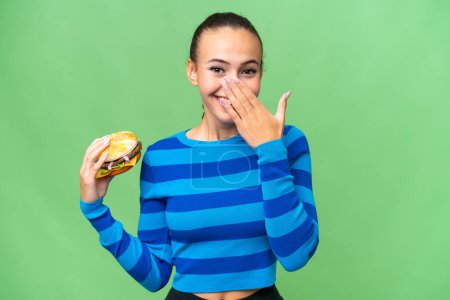 Foto de Mujer árabe joven sosteniendo una hamburguesa sobre un fondo aislado feliz y sonriente cubriendo la boca con la mano - Imagen libre de derechos