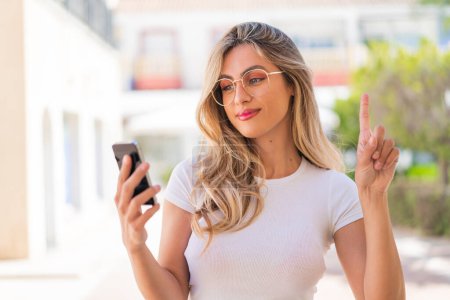 Hübsche blonde uruguayische Frau benutzt Handy und hebt den Finger
