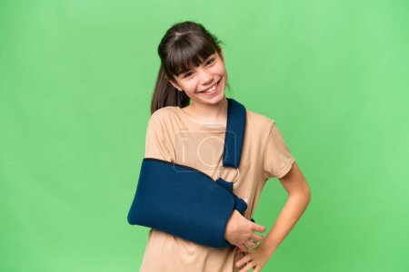 Photo for Little caucasian girl with broken arm and wearing a sling over isolated background posing with arms at hip and smiling - Royalty Free Image