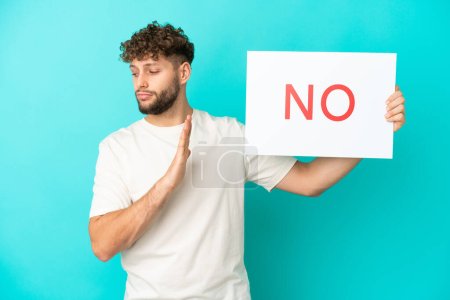Young handsome caucasian man isolated on blue background holding a placard with text NO and doing stop sign
