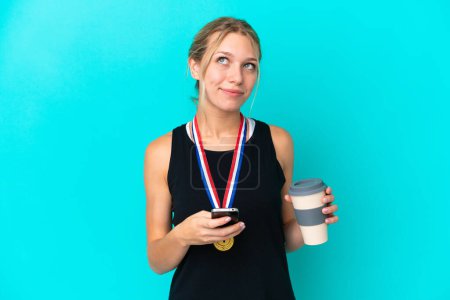 Photo for Young caucasian woman with medals isolated on blue background holding coffee to take away and a mobile while thinking something - Royalty Free Image
