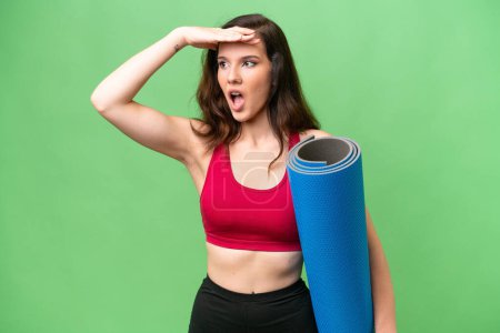 Téléchargez les photos : Jeune femme sportive allant à des cours de yoga tout en tenant un tapis sur fond isolé faisant geste de surprise tout en regardant sur le côté - en image libre de droit