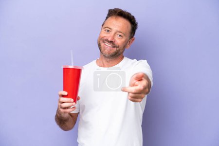Foto de Hombre caucásico de mediana edad sosteniendo soda aislada sobre fondo púrpura apuntando frente con expresión feliz - Imagen libre de derechos