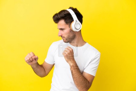 Young caucasian handsome man isolated on yellow background listening music and dancing