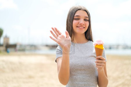 Foto de Chica adolescente con un helado de corneta al aire libre saludando con la mano con expresión feliz - Imagen libre de derechos