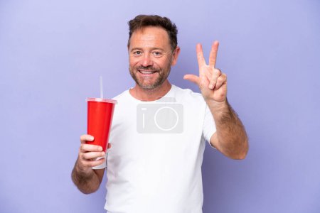 Foto de Hombre caucásico de mediana edad sosteniendo soda aislada sobre fondo morado feliz y contando tres con los dedos - Imagen libre de derechos
