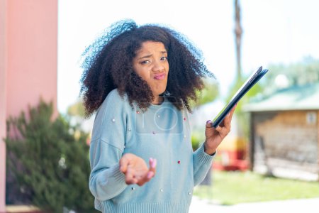 Joven mujer afroamericana sosteniendo una tableta al aire libre haciendo gestos de dudas mientras levanta los hombros