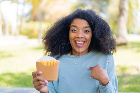 Téléchargez les photos : Jeune femme afro-américaine tenant des chips frites à l'extérieur avec une expression faciale surprise - en image libre de droit
