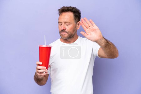 Foto de Hombre caucásico de mediana edad sosteniendo soda aislada sobre fondo púrpura haciendo gesto de parada y decepcionado - Imagen libre de derechos