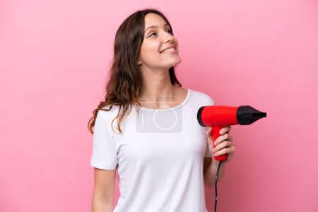 Téléchargez les photos : Jeune femme caucasienne tenant un sèche-cheveux isolé sur fond rose levant les yeux tout en souriant - en image libre de droit