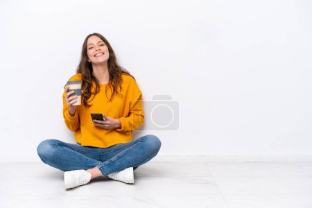 Téléchargez les photos : Jeune femme caucasienne assise sur le sol isolée sur un mur blanc tenant du café à emporter et un mobile - en image libre de droit