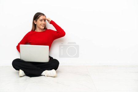 Foto de Joven mujer caucásica con un portátil sentado en el suelo mirando lejos con la mano para mirar algo - Imagen libre de derechos