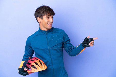 Téléchargez les photos : Jeune cycliste caucasien homme isolé sur fond violet pointant doigt sur le côté et présentant un produit - en image libre de droit