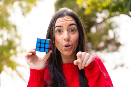 Téléchargez les photos : Jeune femme tenant un cube puzzle tridimensionnel à l'extérieur surpris et pointant devant - en image libre de droit