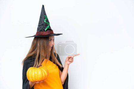 Téléchargez les photos : Jeune costume de femme caucasienne en sorcière tenant une citrouille isolée sur fond blanc pointant du doigt sur le côté et présentant un produit - en image libre de droit