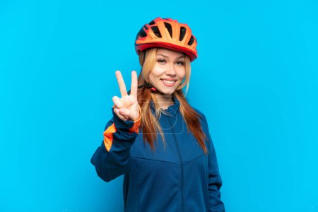 Téléchargez les photos : Jeune cycliste fille isolée sur fond bleu souriant et montrant signe de victoire - en image libre de droit