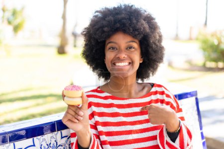Foto de Chica afroamericana sosteniendo una dona al aire libre con expresión facial sorpresa - Imagen libre de derechos