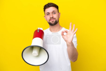 Photo for Young caucasian man isolated on yellow background holding a megaphone and showing ok sign with fingers - Royalty Free Image