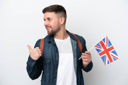 Foto de Joven hombre caucásico sosteniendo una bandera del Reino Unido aislado sobre fondo blanco apuntando hacia un lado para presentar un producto - Imagen libre de derechos