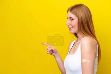 Foto de Mujer pelirroja joven con una tirita aislada sobre fondo amarillo apuntando hacia un lado para presentar un producto - Imagen libre de derechos