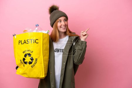 Téléchargez les photos : Jeune femme rousse tenant un sac rempli de bouteilles en plastique à recycler isolé sur fond rose dans le but de réaliser la solution tout en levant le doigt - en image libre de droit
