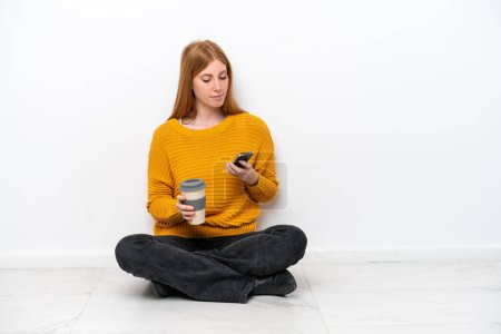 Téléchargez les photos : Jeune rousse femme assise sur le sol isolé sur fond blanc tenant du café à emporter et un mobile - en image libre de droit