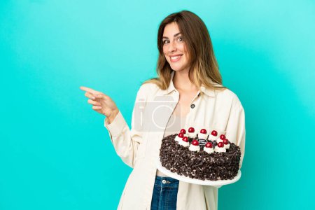 Téléchargez les photos : Femme caucasienne tenant gâteau d'anniversaire isolé sur fond bleu pointant doigt sur le côté - en image libre de droit