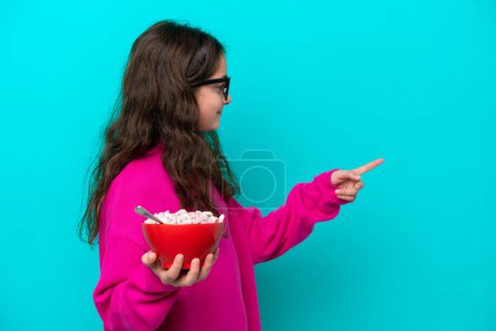 Téléchargez les photos : Petite fille tenant un bol de céréales isolé sur fond bleu pointant vers le côté pour présenter un produit - en image libre de droit