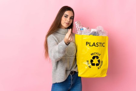 Foto de Joven mujer caucásica sosteniendo una bolsa llena de botellas de plástico para reciclar aislado sobre fondo rosa haciendo gesto de dinero - Imagen libre de derechos